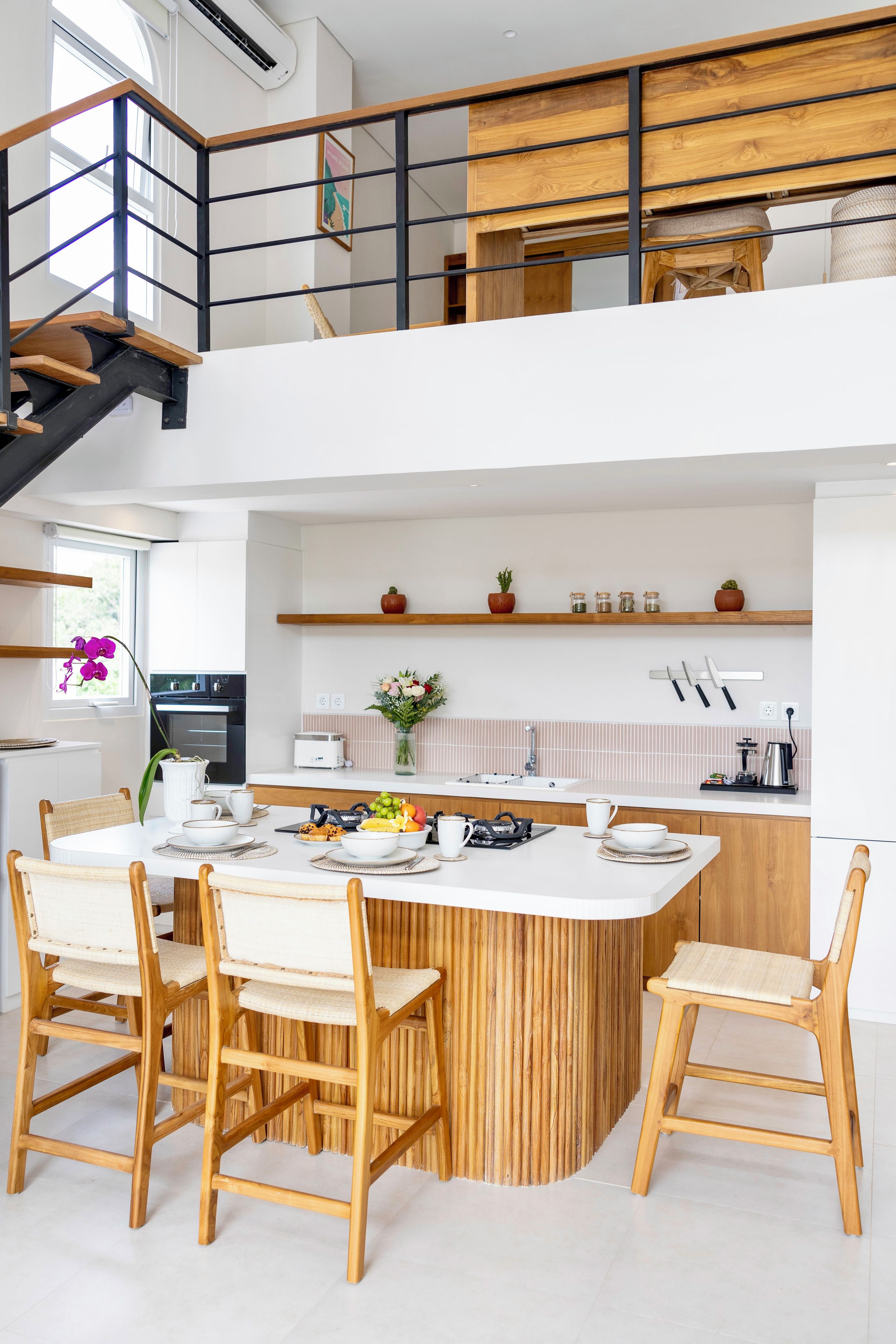 A modern kitchen with a central island, wooden chairs, and a staircase leading to an upper level.