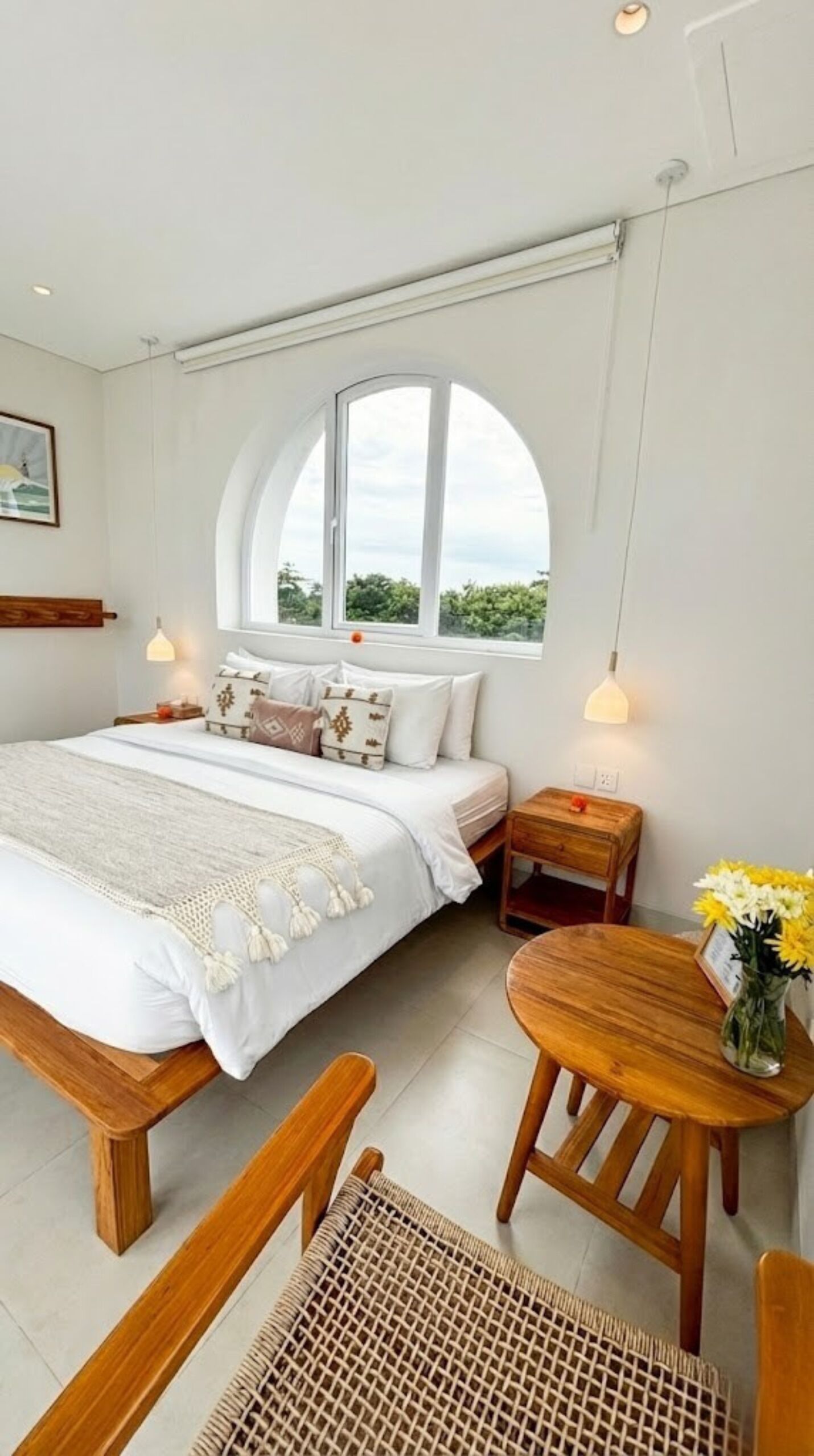 Un dormitorio con una cama grande, un banco de madera, una mesita con un florero y un ventanal con vistas a la vegetación.