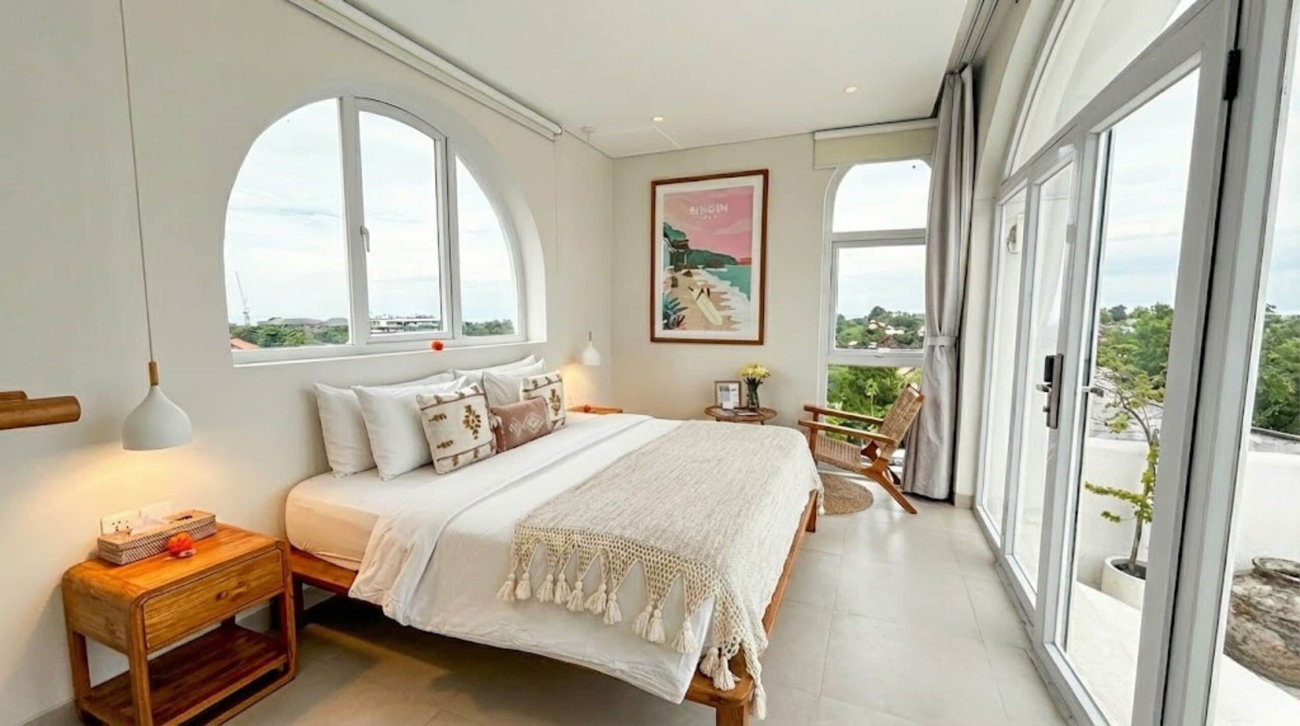 A bedroom with a large bed, a wooden nightstand, a small table with a lamp, and a chair. There are large windows with a view of greenery outside.