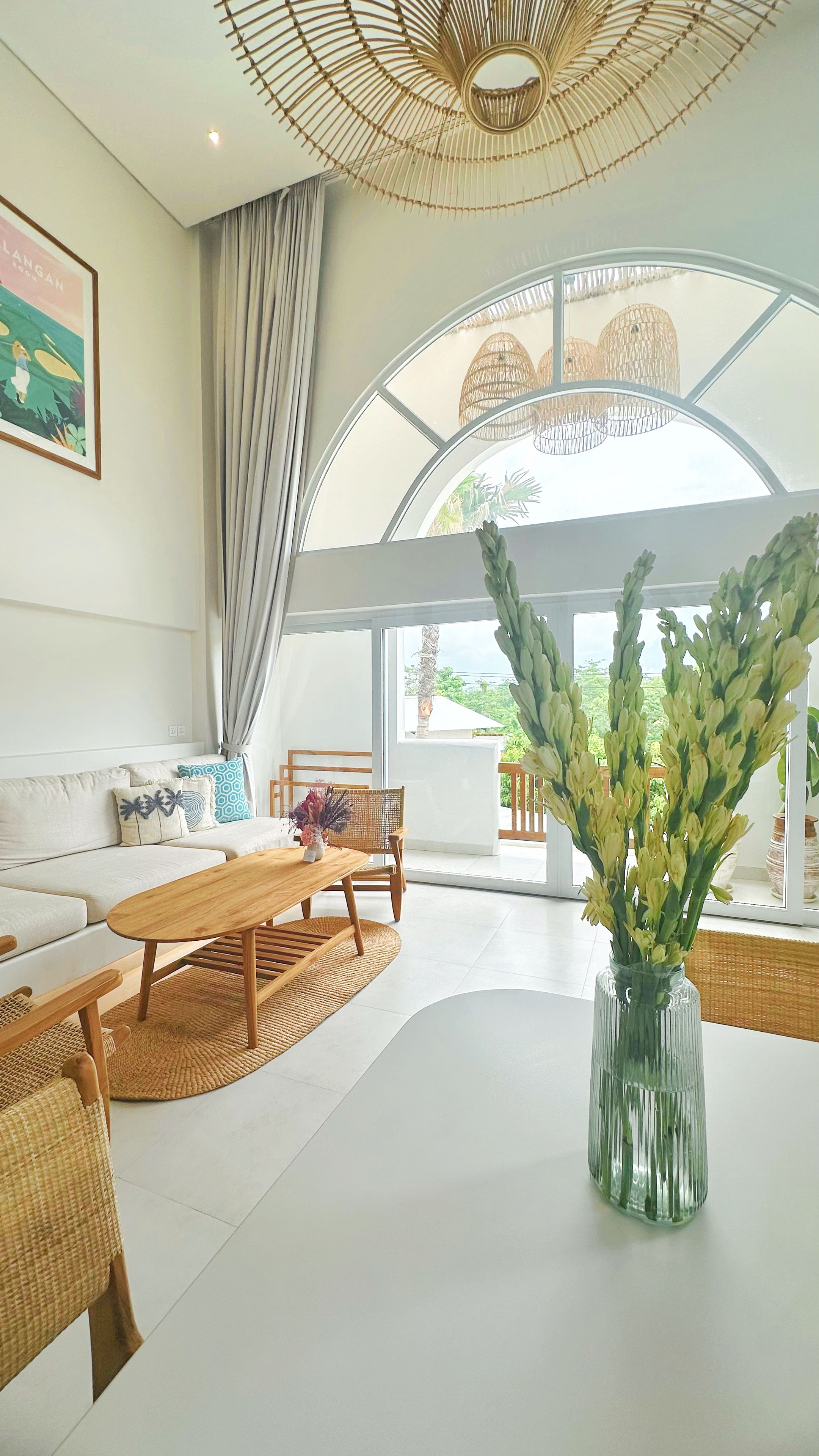 A spacious living area with a sofa, wooden coffee table, and wicker chairs. A large window offers a view of greenery outside.