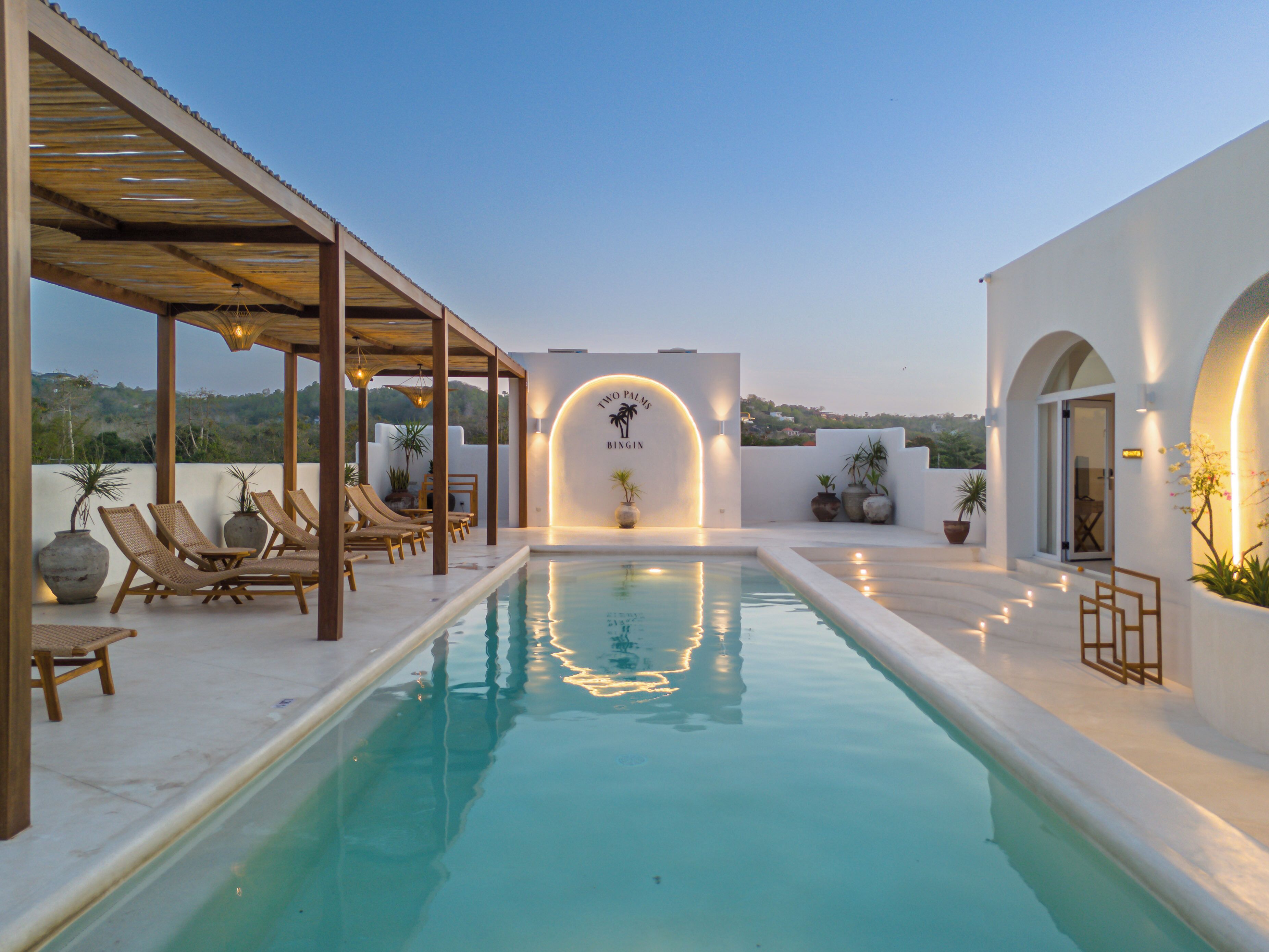 A pool area with lounge chairs, a pergola, and a wall with a palm tree logo.