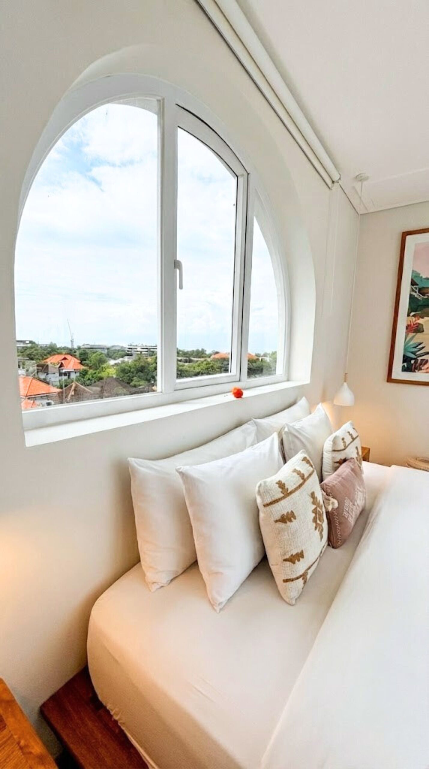 A bedroom with a large arched window, a bed with white bedding, and a view of buildings and greenery outside.