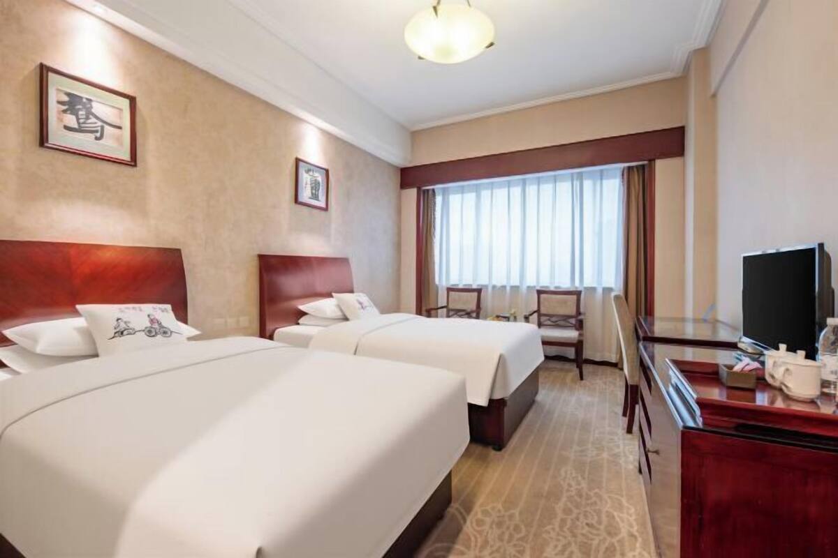 Quarto de hotel com duas camas, uma escrivaninha, televisão e uma janela grande com cortinas.