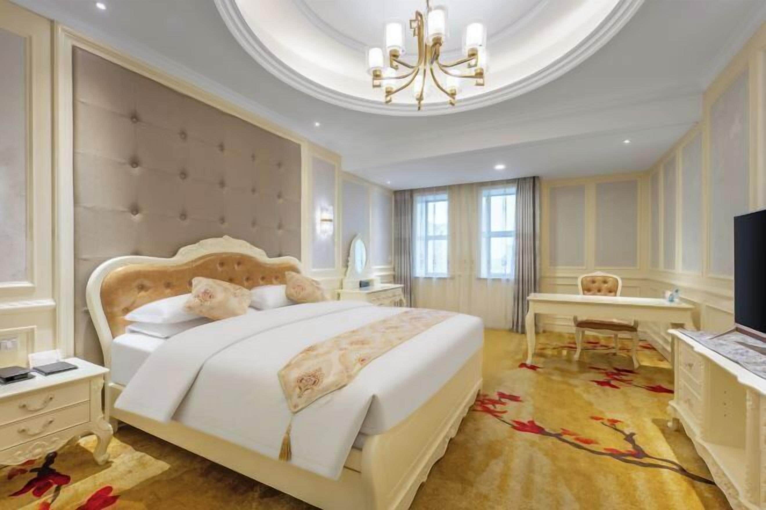 A hotel room with a large bed, a desk, a chair, a television, and a chandelier.