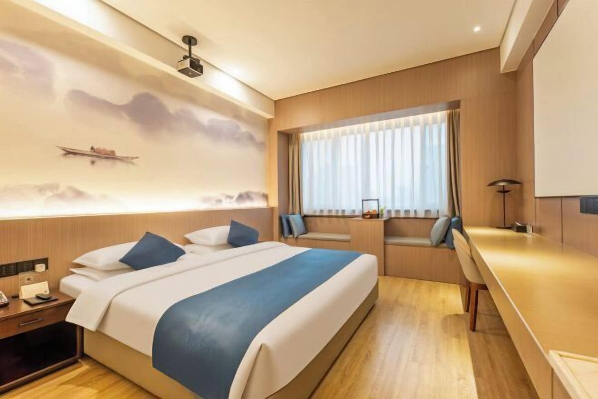 Quarto de hotel com cama, mesa de cabeceira, luminária, janela com cortinas e prateleira na parede.