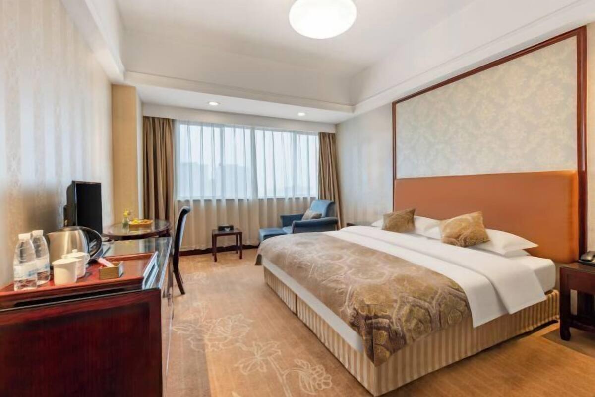 Quarto de hotel com uma cama grande, uma escrivaninha com cadeira, uma televisão e uma janela com cortinas.
