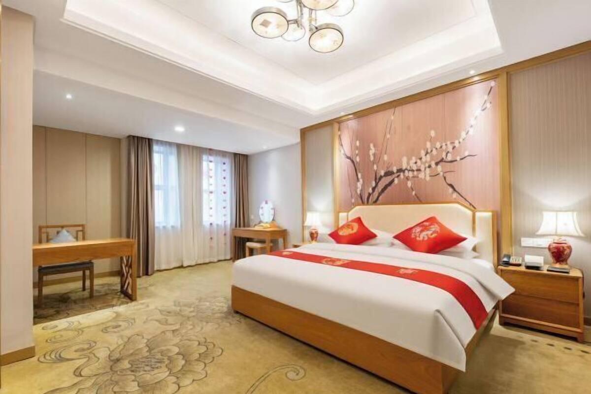 Quarto de hotel com uma cama grande, uma escrivaninha, uma cadeira, uma mesa de cabeceira e um painel decorativo na parede.