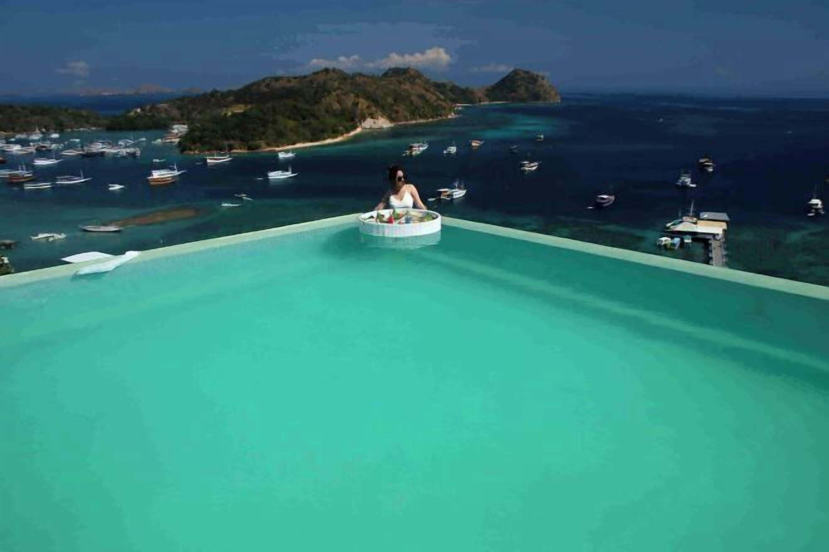 Uma pessoa em uma plataforma circular flutuante em uma piscina azul infinita, com vista para barcos e uma ilha distante.