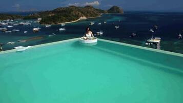 Uma pessoa em uma plataforma circular flutuante em uma piscina azul infinita, com vista para barcos e uma ilha distante.
