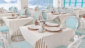 A restaurant with tables set for dining, chairs with patterned cushions, and a view of the sea through large windows. - Loccal Collection Hotel Komodo (Labuan Bajo)