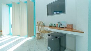 A room with a large window, a TV mounted on the wall, a wooden desk with various items, a wicker chair, and a small black refrigerator. - Loccal Collection Hotel Komodo (Labuan Bajo)