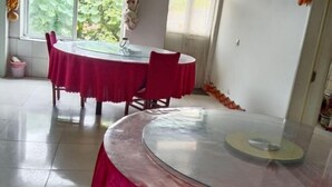 Uma mesa de vidro redonda com toalha de mesa vermelha, rodeada por cadeiras, em um quarto com piso de azulejos e uma janela com vegetação lá fora.