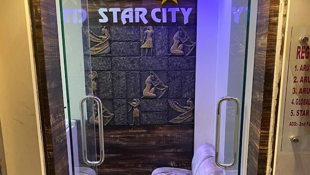 A hotel room with a glass door, a bed with patterned bedding, and a wall with Egyptian-themed artwork. - Hotel Star City (Kolkata)