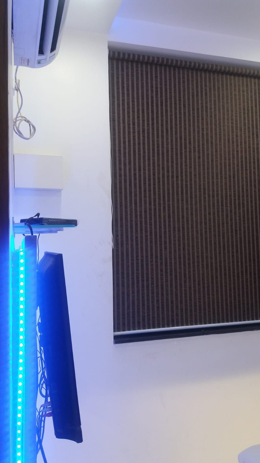 A room with a large window covered by vertical blinds, a white air conditioning unit, and a blue LED light strip on the left side.