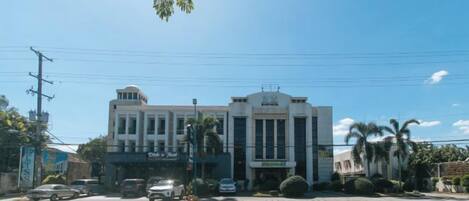 A multi-story building with a central entrance, a parking lot in front, and palm trees in the background.