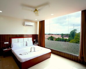A hotel room with a large bed, an air conditioning unit, a nightstand, a telephone, and a view of the city through a large window. - Hotel Vishnu Inn (Dehradun)