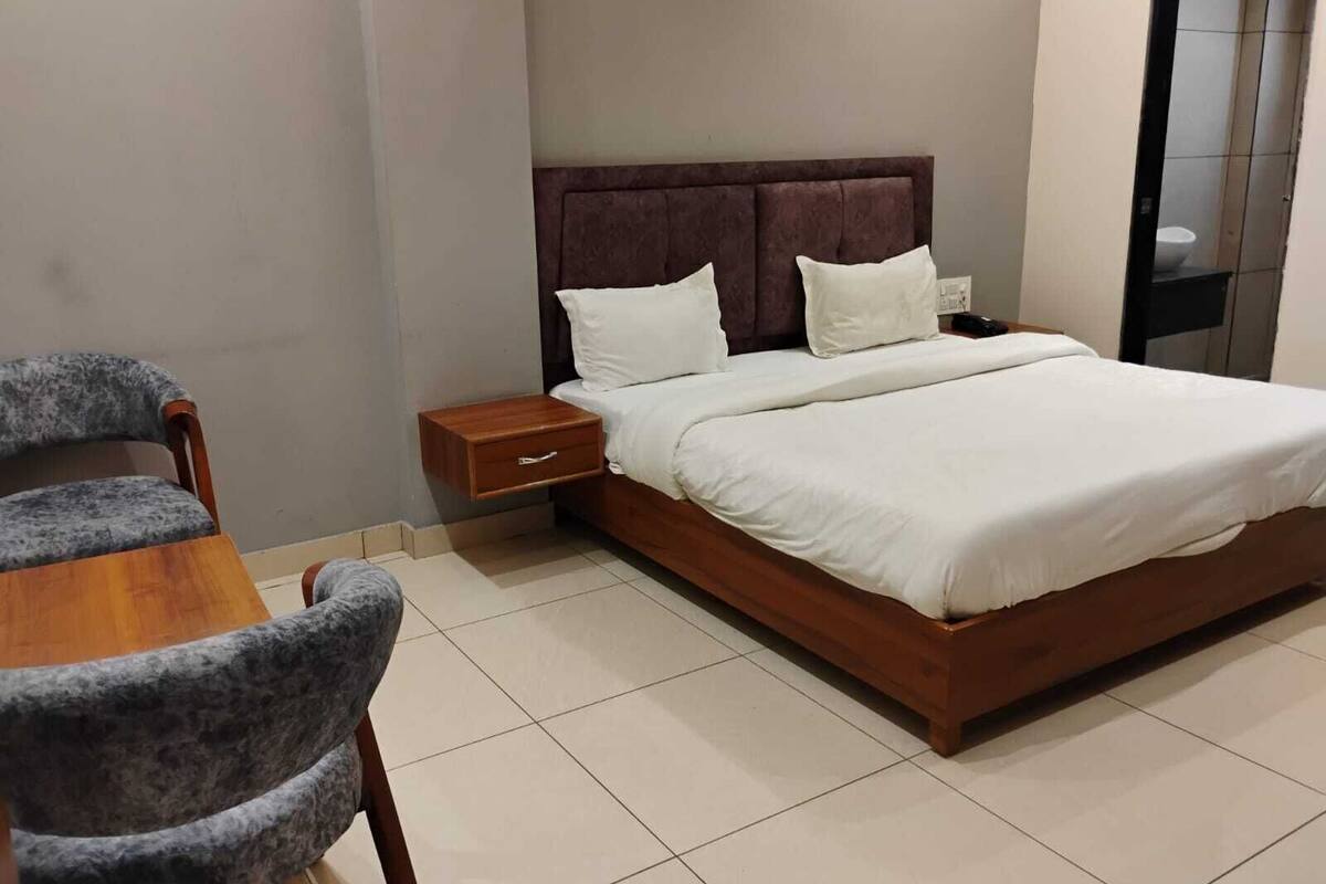 A neatly made bed with white linens, a wooden nightstand with a drawer, and a grey upholstered chair.
