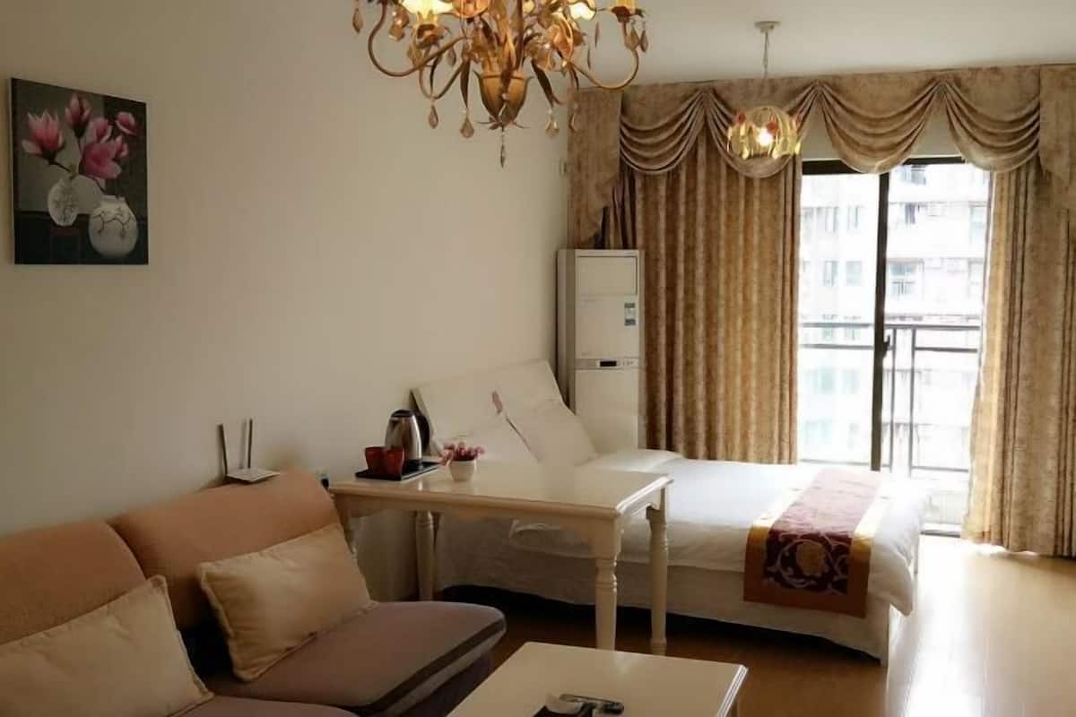 A hotel room with a bed, a sofa, a table, a chandelier, a painting, and a window with curtains.
