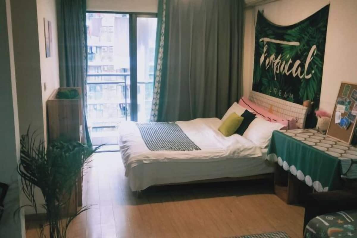 A bedroom with a bed, a window with curtains, a green plant, and a wall with a tropical-themed poster.