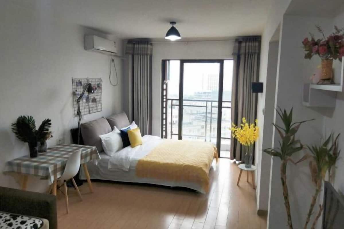 A bedroom with a bed, a desk, a chair, a window with a view, and a potted plant.