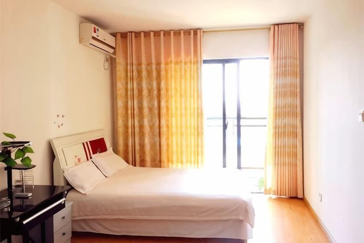 Une chambre à coucher avec un lit, une table de chevet, une plante et une fenêtre avec des rideaux.