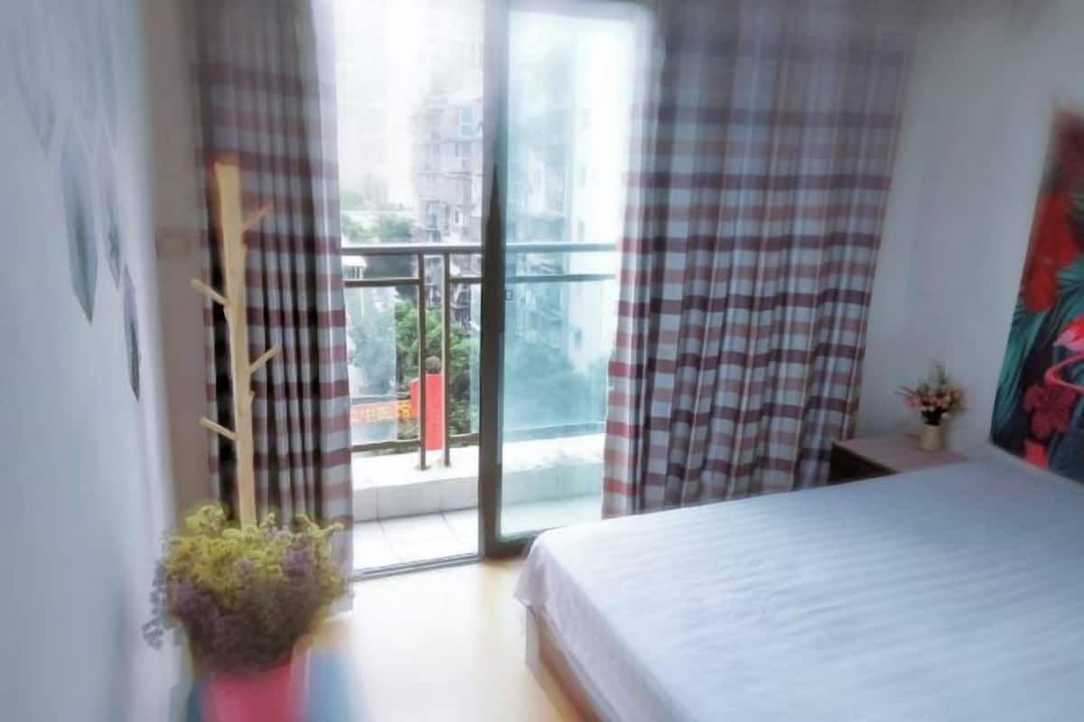 A bedroom with a bed, a potted plant, a window with curtains, and a view of buildings outside.