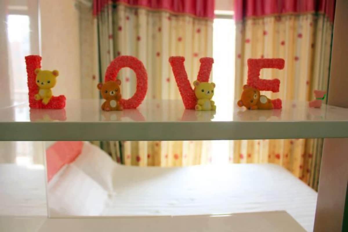 Ein Bett mit weißer Matratze und Kissen, ein Glastischchen mit Teddybären und der Schriftzug 'LOVE' in roten Buchstaben, sowie ein Fenster mit rosa Vorhängen im Hintergrund.