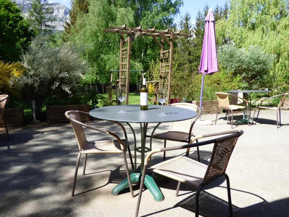 An outdoor patio with a round table and four wicker chairs, surrounded by greenery and a wooden pergola.