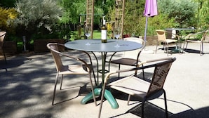 Un patio exterior con una mesa redonda y cuatro sillas de mimbre, rodeado de follaje y una pérgola de madera.