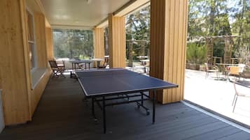 An outdoor wooden deck with a ping pong table and chairs, surrounded by wooden walls and large windows.