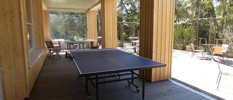 Terraza exterior de madera con mesa de ping pong y sillas, rodeada de paredes de madera y grandes ventanales.