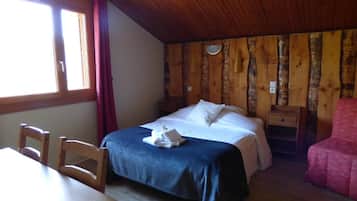 A bedroom with a wooden ceiling, a bed with white linens, a wooden nightstand, a table with chairs, and a window with curtains.
