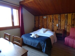 Un dormitorio con techo de madera, una cama con ropa blanca, una mesita de noche de madera, una mesa con sillas y una ventana con cortinas.