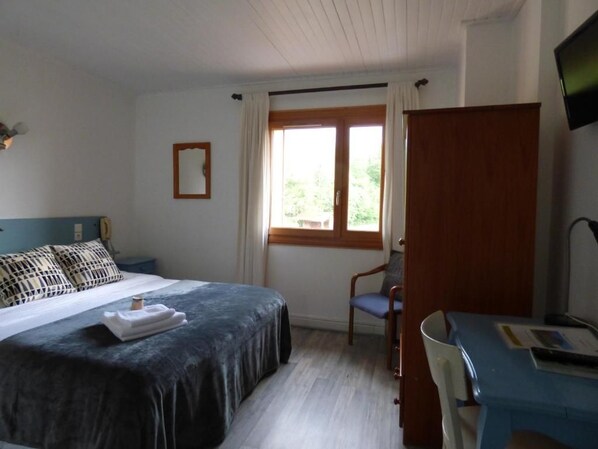 A bedroom with a bed, a desk, a chair, a window, and a TV.