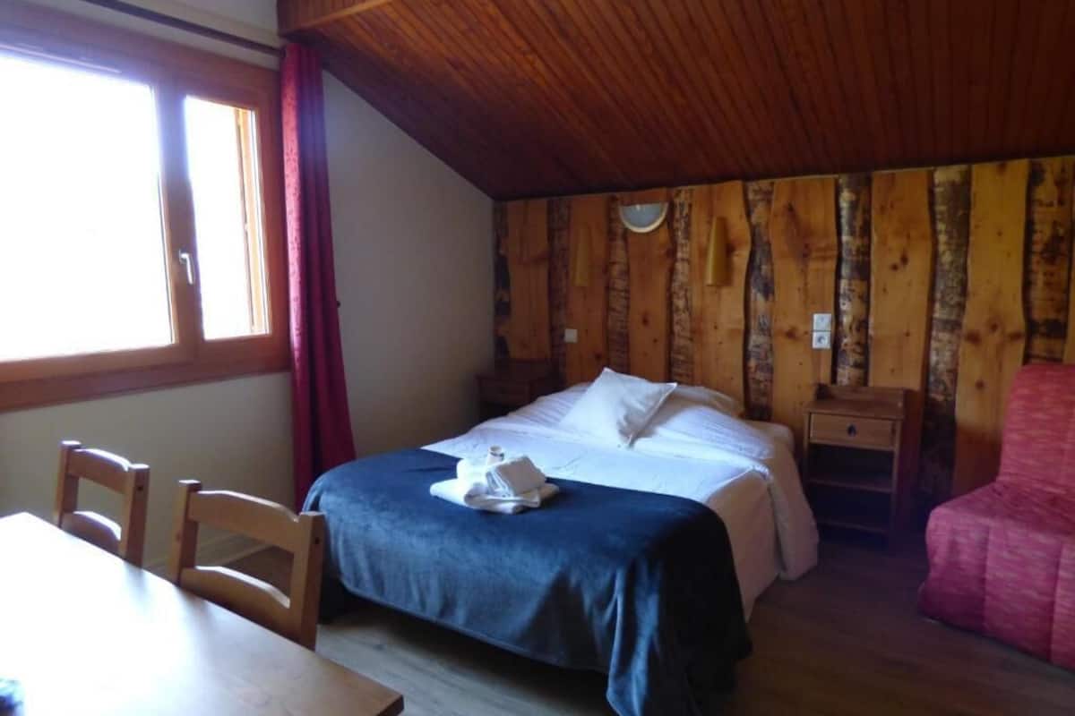 A bedroom with a wooden ceiling, a bed with white linens, a wooden nightstand, a table with chairs, and a window with curtains.