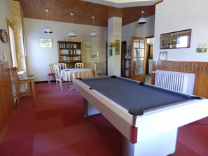Mesa de billar en una habitación con alfombra roja, paredes y techo de madera, rodeada de sillas y una estantería.