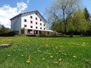 A multi-story building with a white facade and brown window frames, surrounded by a lush green lawn with yellow flowers. - Logis Hotel La Cremaillere St Bonnet (Saint-Bonnet-en-Champsaur)