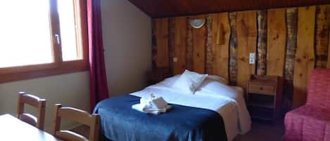 A bedroom with a wooden ceiling, a bed with white linens, a wooden nightstand, a table with chairs, and a window with curtains.