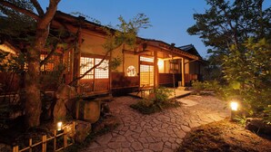 Une maison traditionnelle japonaise, avec un chemin de pierres menant à l’entrée, entourée d’arbres et d’un petit jardin.