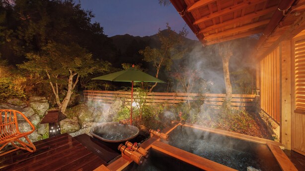 Un espace de baignade dans une source thermale, avec des bancs en bois et un parasol vert, entouré d’un paysage naturel et de constructions en bois.