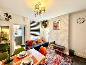 En moderne stue med spiseplads, en sofa med puder, et lille sofabord, et ur på væggen og en reol.