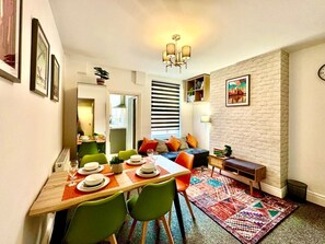 A dining area with a wooden table set for four, green chairs, a colorful rug, a sofa with orange cushions, and a bookshelf.