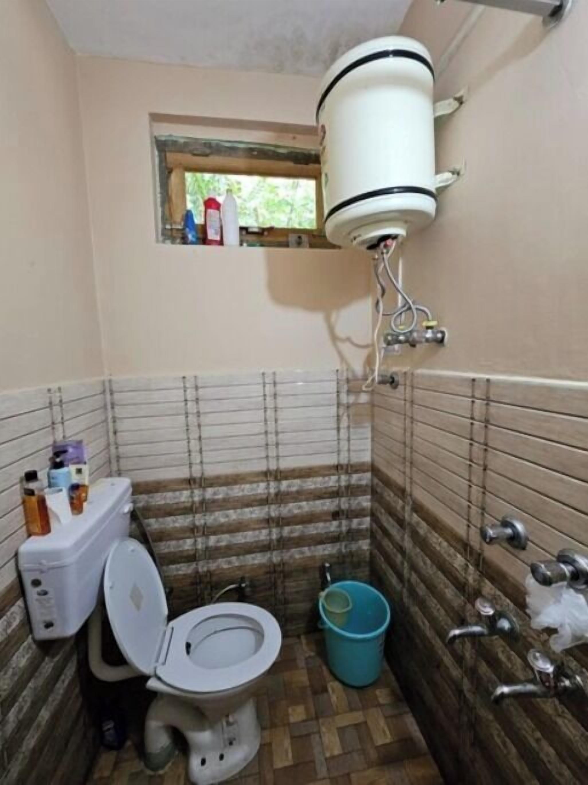 A small bathroom with a toilet, a bidet, a bucket, and a window above the toilet.