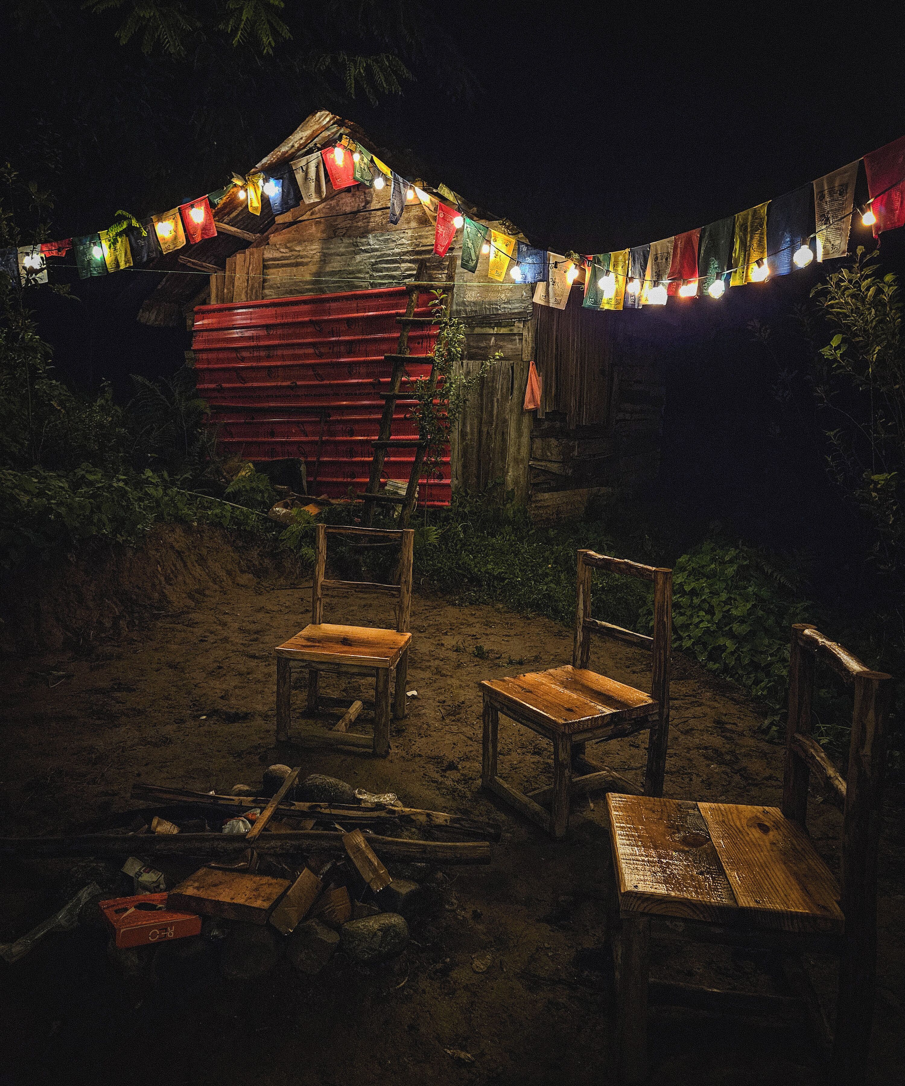 A rustic outdoor setting with wooden chairs and a red wooden structure, possibly a shed or cabin, under a string of colorful lights.