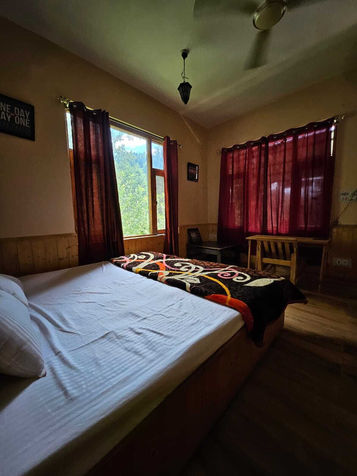 A bedroom with a bed, a desk, and a chair. There is a window with curtains, a ceiling fan, and a sign on the wall.