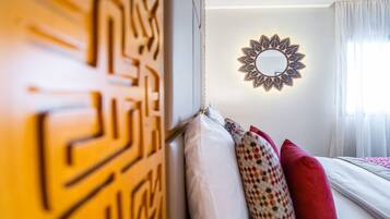 Una habitación de hotel con un diseño geométrico en la pared, un espejo redondo con un motivo similar al sol, y una cama con almohadas blancas y rojas.