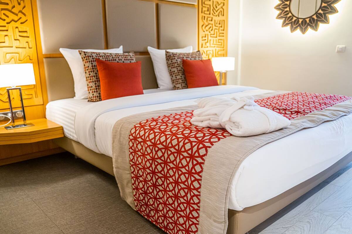 A hotel room with a neatly made bed, a bedside table with a lamp, and a decorative wall panel.