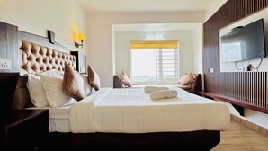 A hotel room with a large bed, a TV mounted on the wall, and a window with blinds. - Kanavu Valley View Resort (Coonoor)