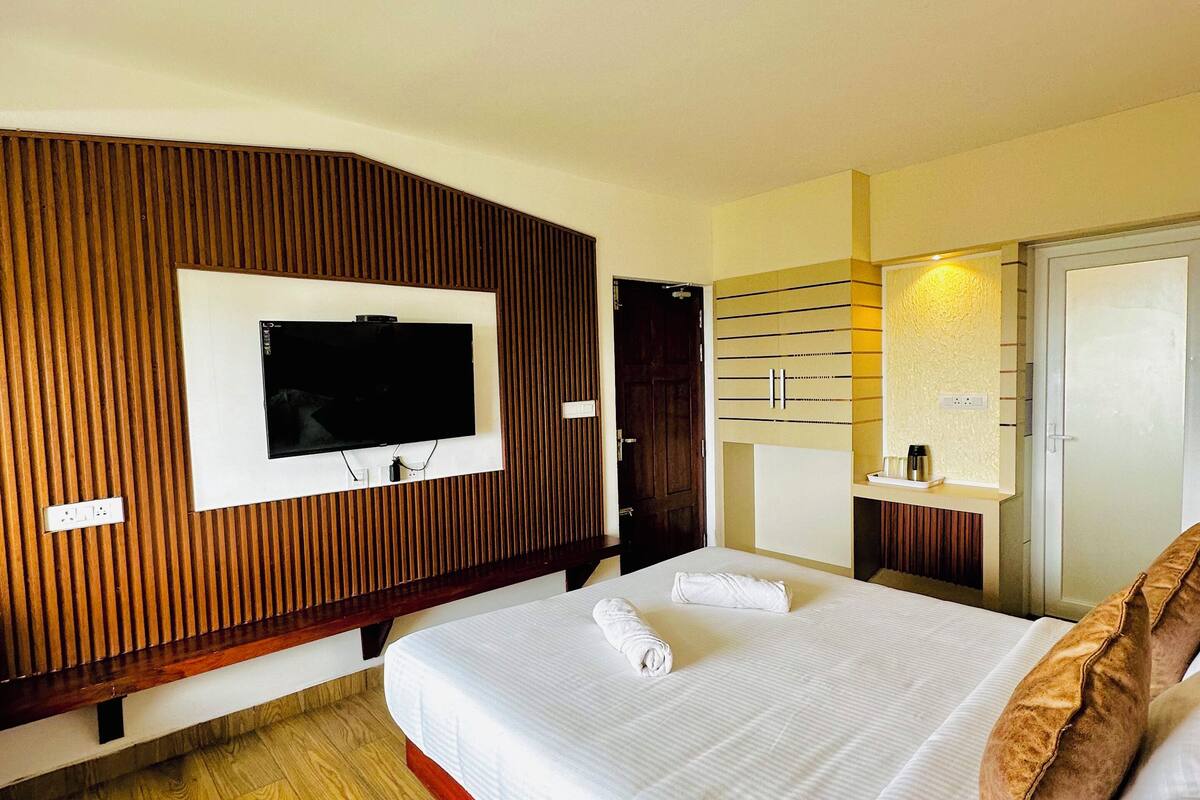 A hotel room with a large bed, a TV mounted on the wall, wooden paneling, a wardrobe, and a small table with a lamp.