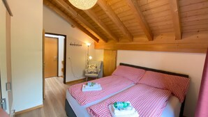 Een slaapkamer met een houten plafond, een bed met rood-wit geruit beddengoed, een stoel en een wandlamp.